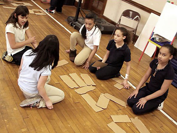 Five young girls are kneeling on a light brown wooden floor, arranging rectangular pieces of cardboard in a grid pattern. One girl in a white shirt kneels beside them, while three others wear black shirts. A fourth girl in a black shirt kneels to the right. A white board and a chair are visible in the background.