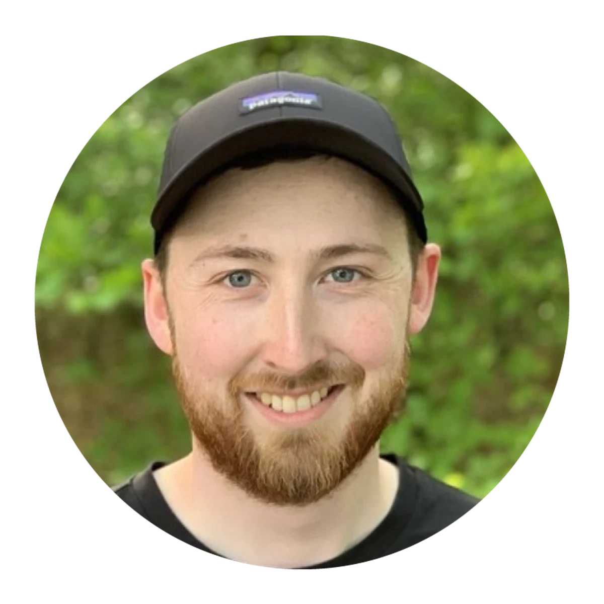 A person outdoors wears a black cap and black top. Green foliage fills the background.