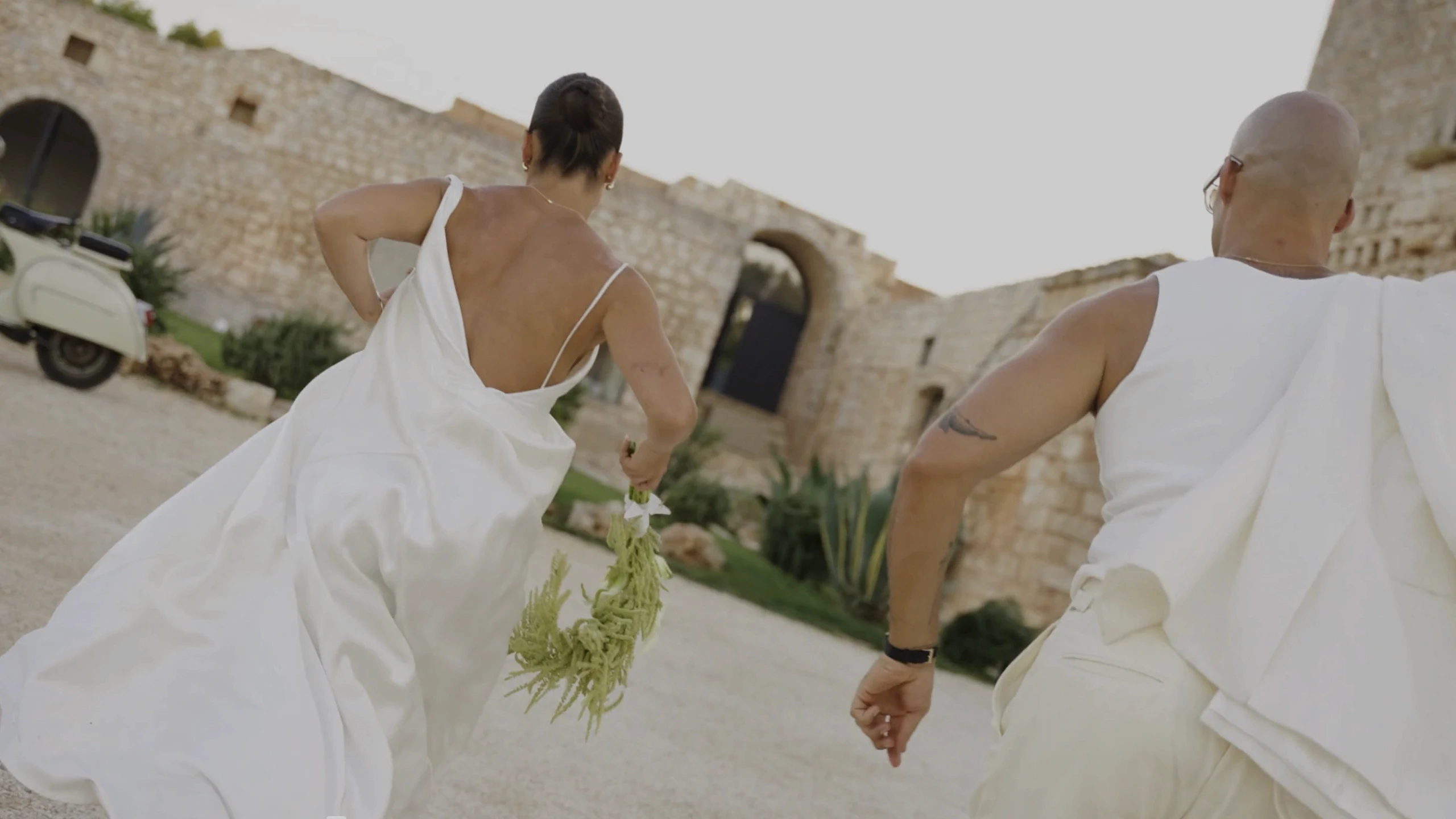 A couple in elegant white attire joyfully runs hand-in-hand outside a rustic stone building, with the bride holding a bouquet of greenery, capturing the essence of a romantic and adventurous wedding escape.