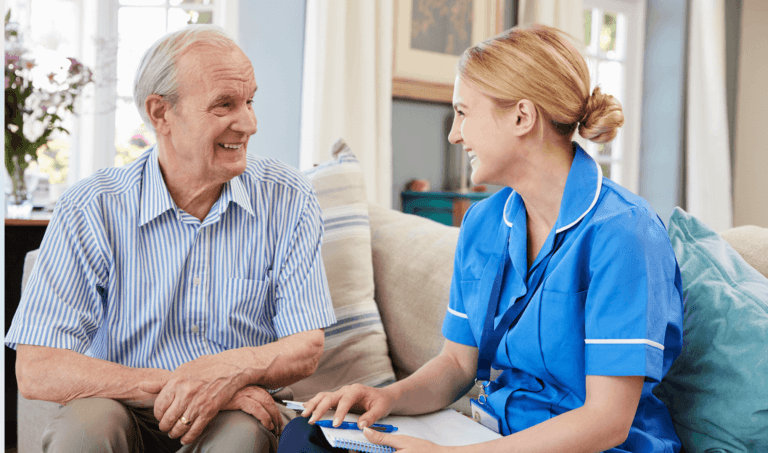 A community nurse in blue scrubs sitting and having a caring conversation with an elderly male patient in a home or care setting