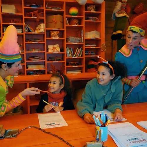 Zwei Kinder an einem Werkstatttisch lächeln zwei Erwachsenen zu, die als Elfen verkleidet sind, in einem bunten, festlichen Raum mit ordentlichen Regalen.