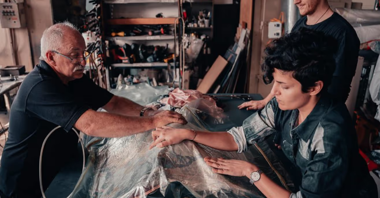 Two people focus on measuring and cutting fabric on a table in a workshop, with various tools and materials in the background. A third person observes the process, standing nearby.