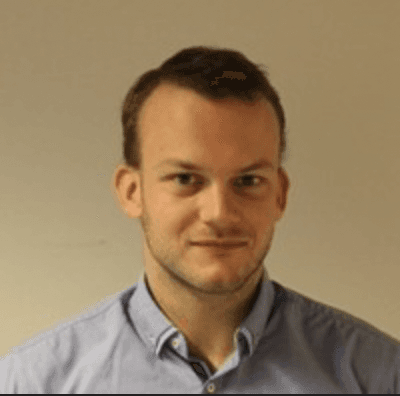 A smiling person with long, curly hair stands with arms crossed, in a modern, blurred background.