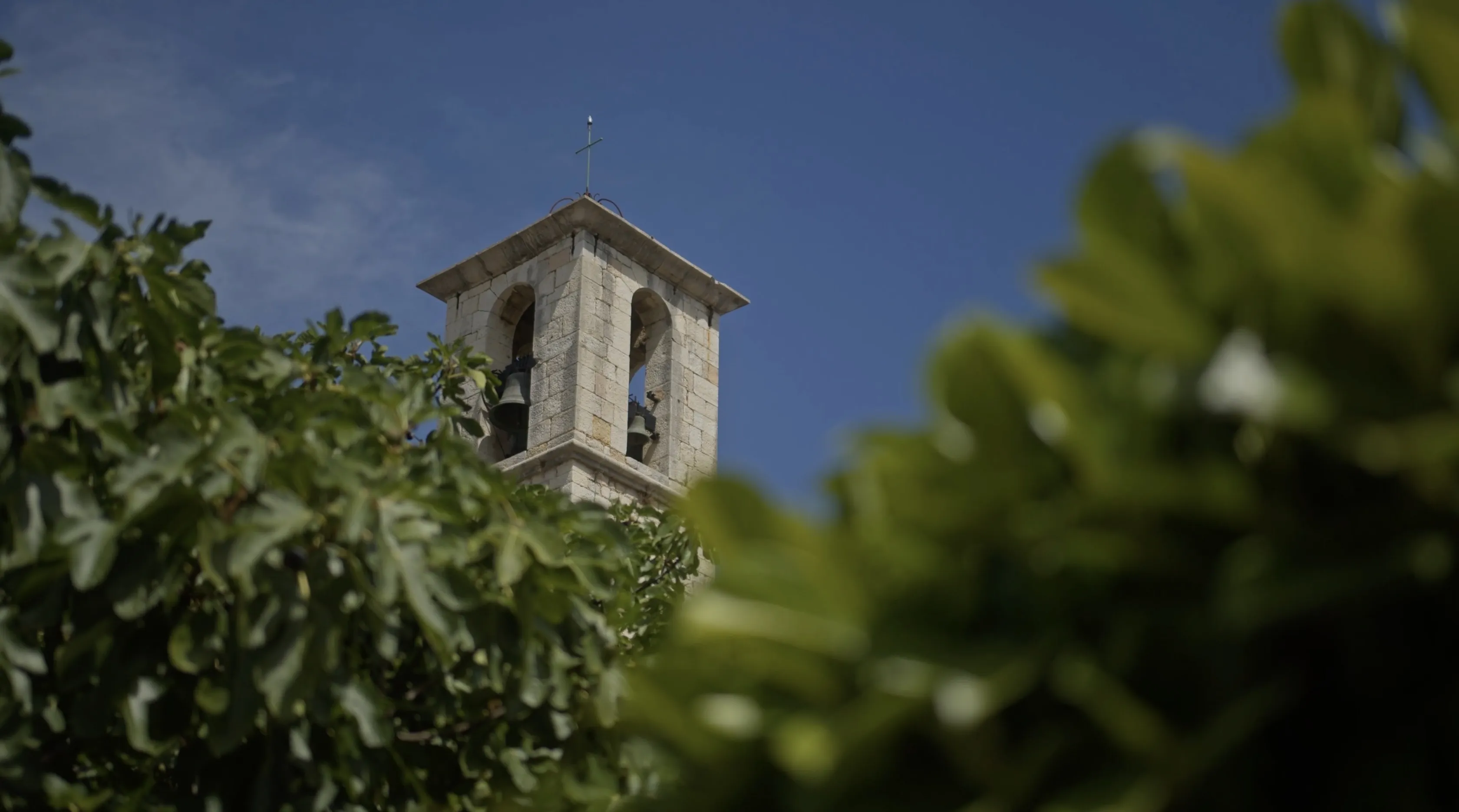 A tall, stone bell tower with visible arches rises above lush green foliage against a clear blue sky.