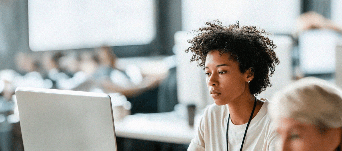 A young person with short, curly hair sits at a desk working on a computer in a bright classroom or office setting. They appear focused on the screen, with other people and desks softly blurred in the background, suggesting a collaborative learning or work environment.