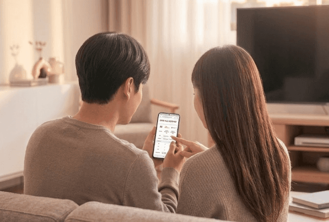 거실 소파에 나란히 앉아 스마트폰 화면으로 2인가구 평균 인터넷 요금을 함께 살펴보며 손가락으로 가리키고 있는 2인 가구 남녀의 뒷모습입니다.