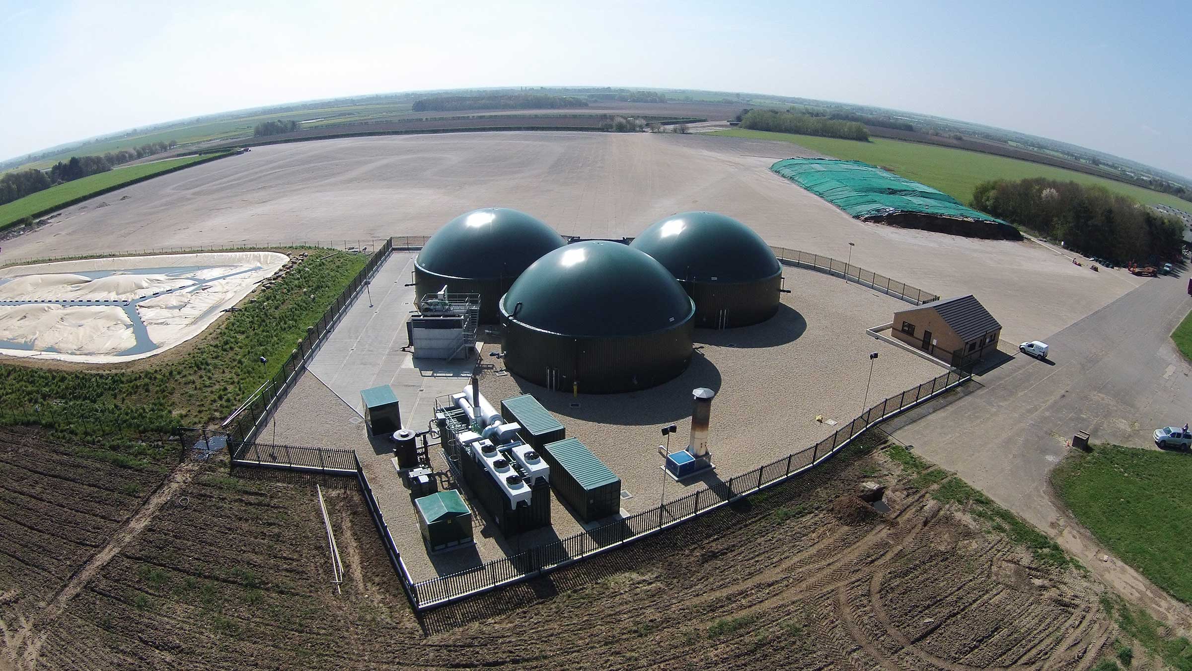 Wide-angle aerial view of anaerobic digestion plant with three green digesters, processing equipment, and control building surrounded by farmland