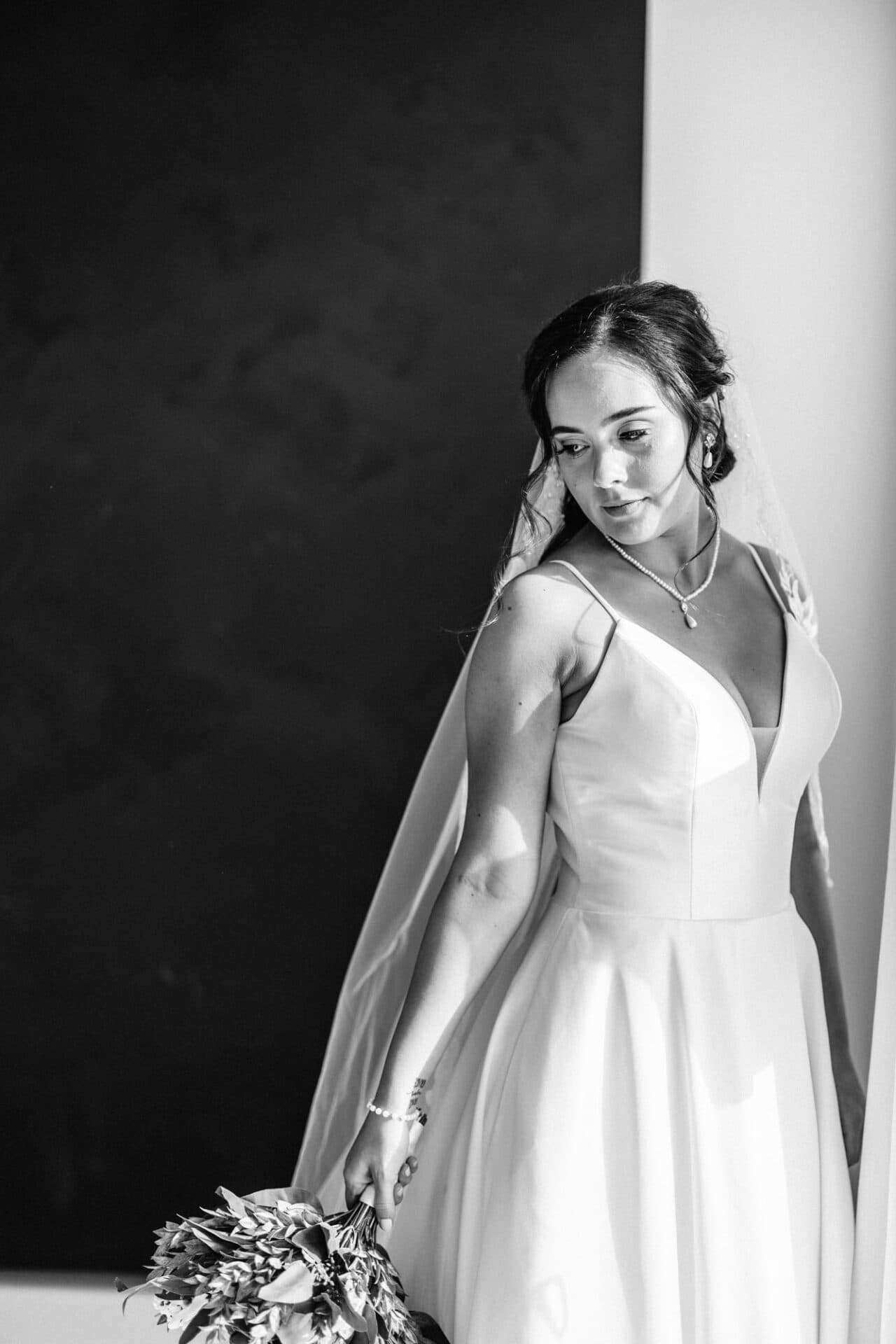 A bride gazes down while standing in a soft-lit room, holding her bouquet at Revelator Studio, a Shreveport natural light photography studio.