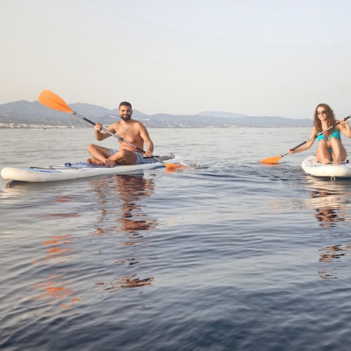 Two people paddleboarding on calm water with mountains in the background, both holding orange paddles and smiling.