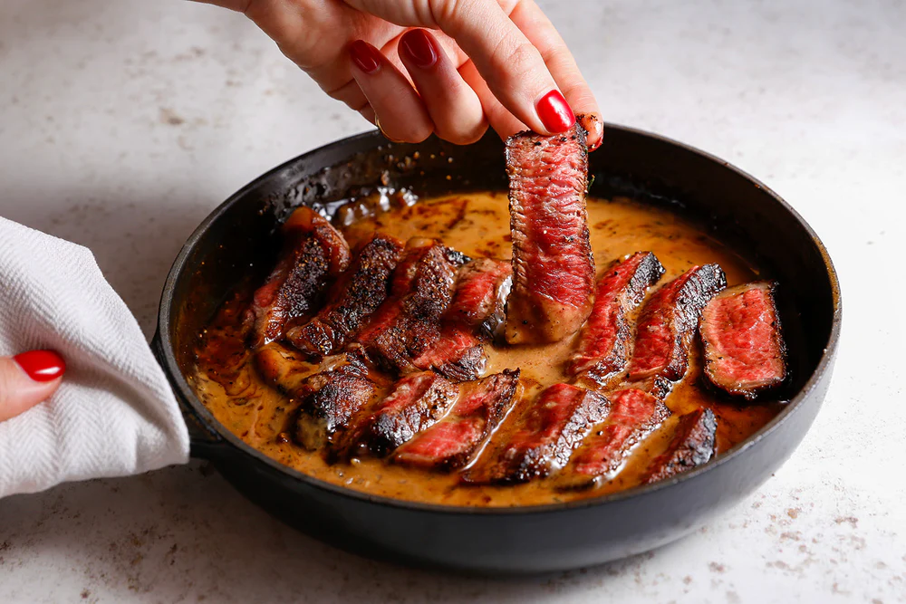 Sliced medium-rare steak in a creamy sauce in a pan, with a hand picking up a piece. The steak has a seared crust and pink center, creating a rich, indulgent food scene.