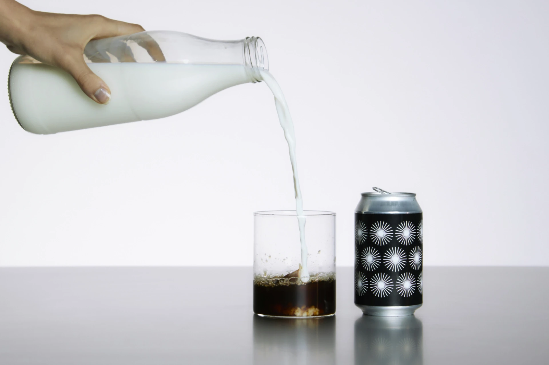 Milch wird in ein Glas mit Cola gegossen, daneben eine Dose mit grafischem Schwarz-Weiß-Muster – Sinnbild für Markenstruktur, visuelle Klarheit und systemische Identität.