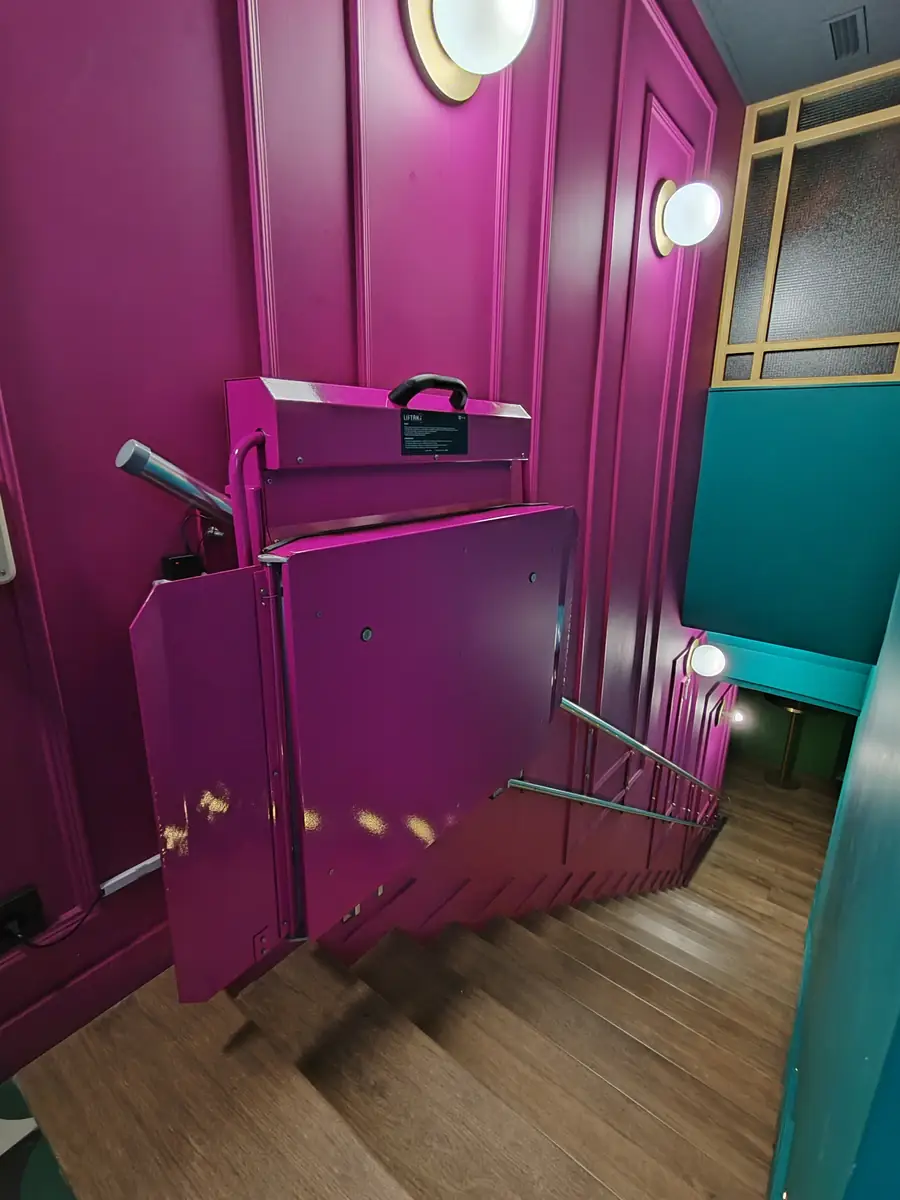 Liftak incline platform stairlift finished in a custom magenta colour, folded against a matching panelled wall in a commercial interior with teal and gold decor