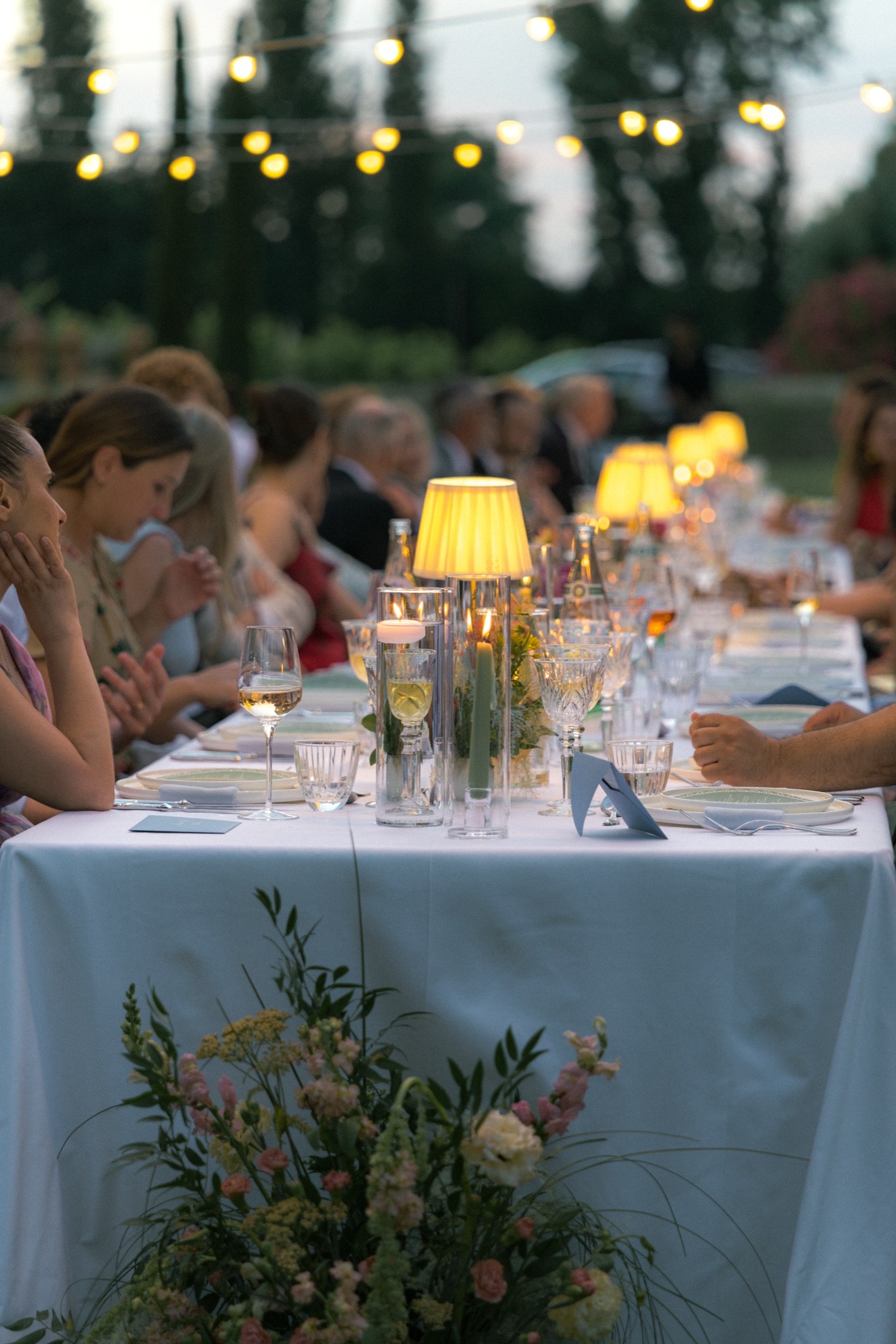 Guests seated along a candlelit dinner table with lamps and flowers