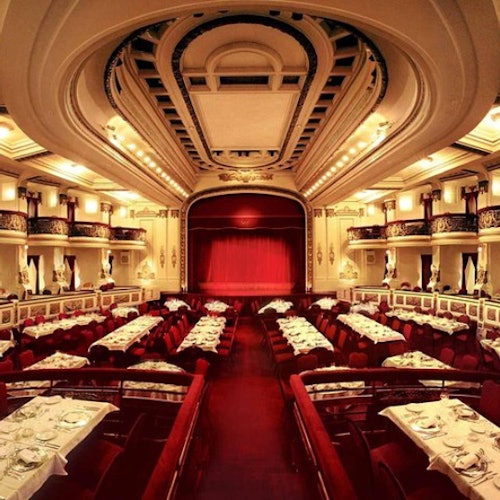 Elegant theater with a stage featuring a red curtain, surrounded by dining tables set with white tablecloths and place settings.