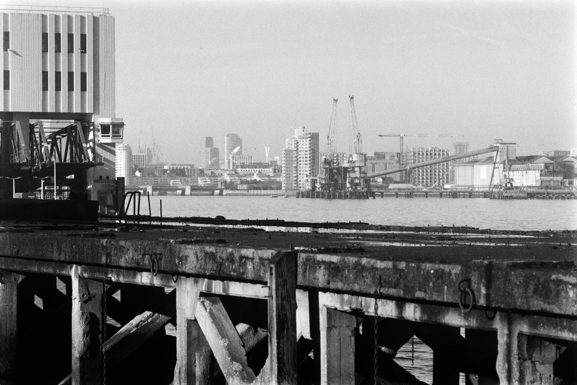Weathered concrete pier with vertical timber supports extending across Thames, corrugated industrial building at left, construction cranes and mid-rise blocks across the water