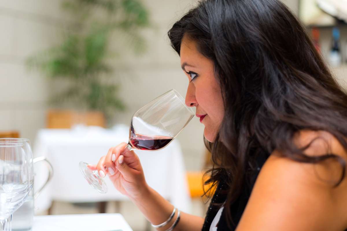 A woman with long dark hair holds a glass of red wine and takes a sip at an outdoor cafe table.