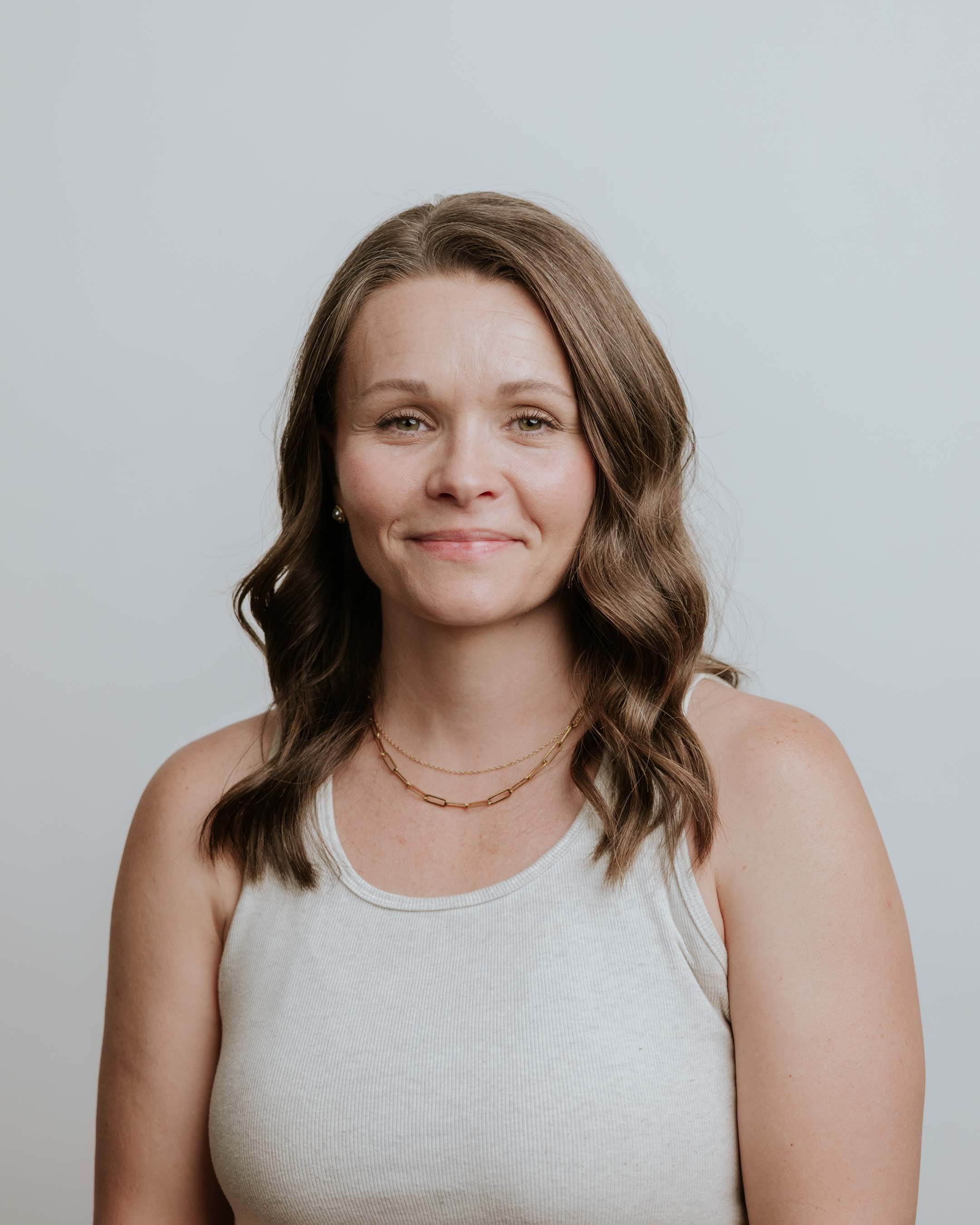 A smiling woman with long brown hair, wearing a brown top, against a plain white background.