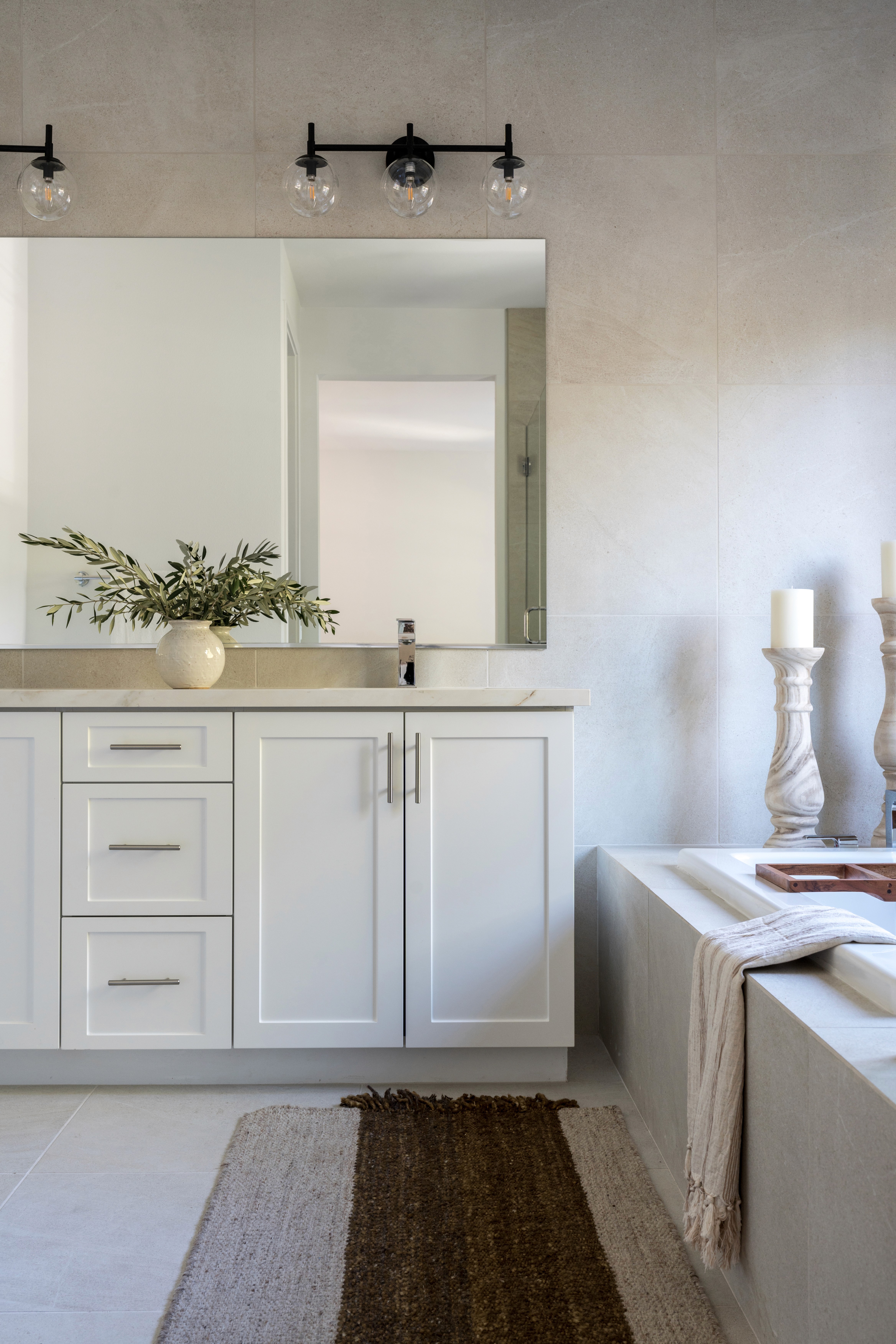 Bright full bath with white cabinets, dark, globe sconces for vanity lighting, and neutral vases and candle stands by Jubilee Interiors in Arcadia, California