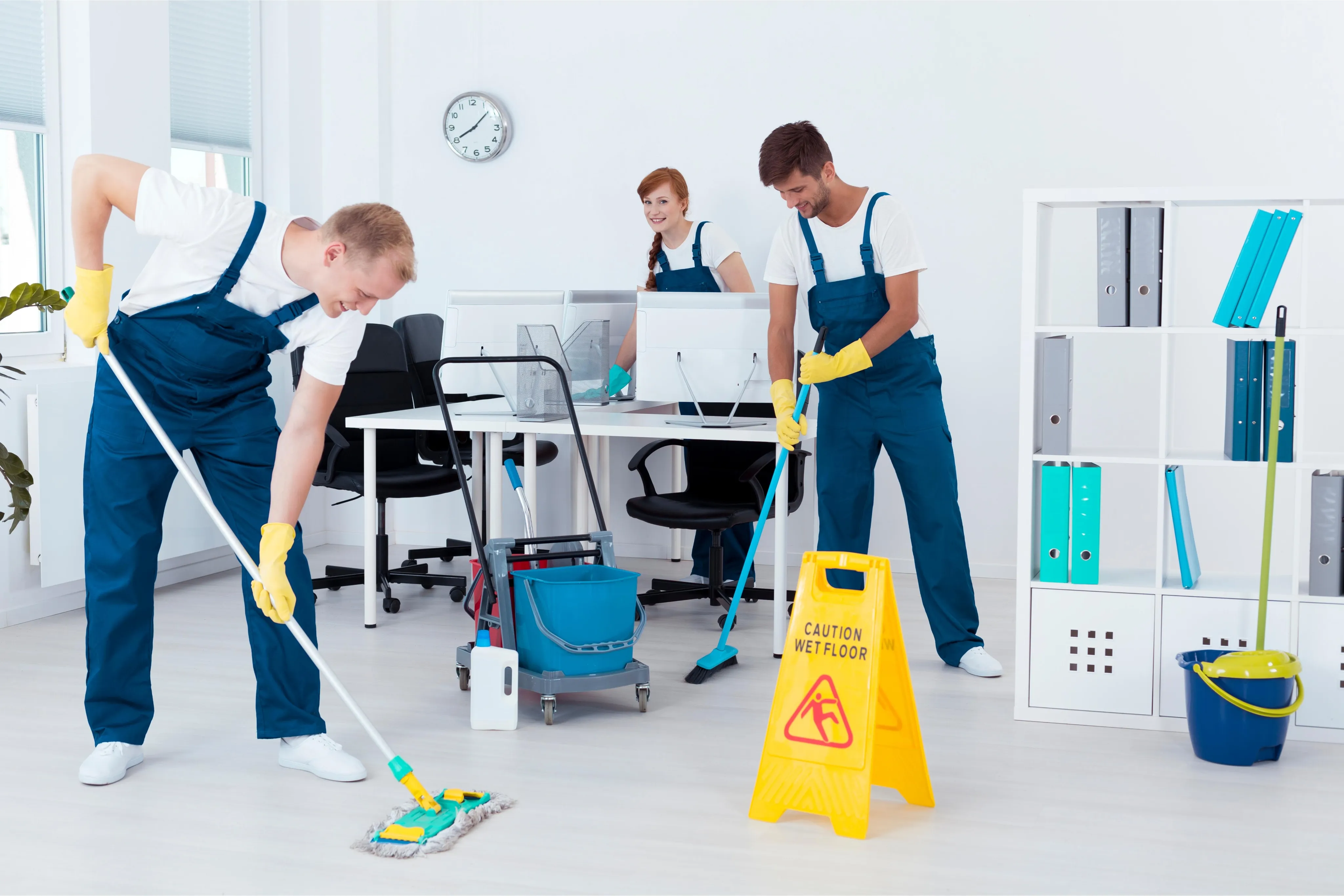 Three professional cleaners in blue overalls and yellow gloves performing deep cleaning services in a modern, bright office space with a "Caution Wet Floor" sign and cleaning cart.