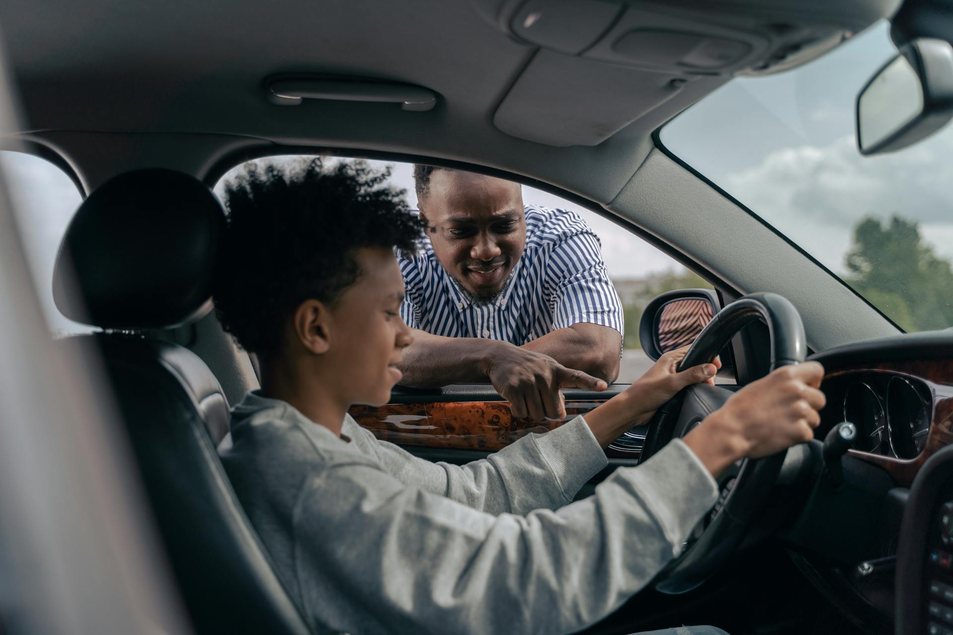 A young person with curly hair, wearing a grey sweatshirt, is seated in the driver's seat of a car, holding the steering wheel. An older man, wearing a striped shirt, is leaning down from outside the car, looking in through the open driver's window and smiling, while pointing toward the front of the car, seemingly giving driving instructions.