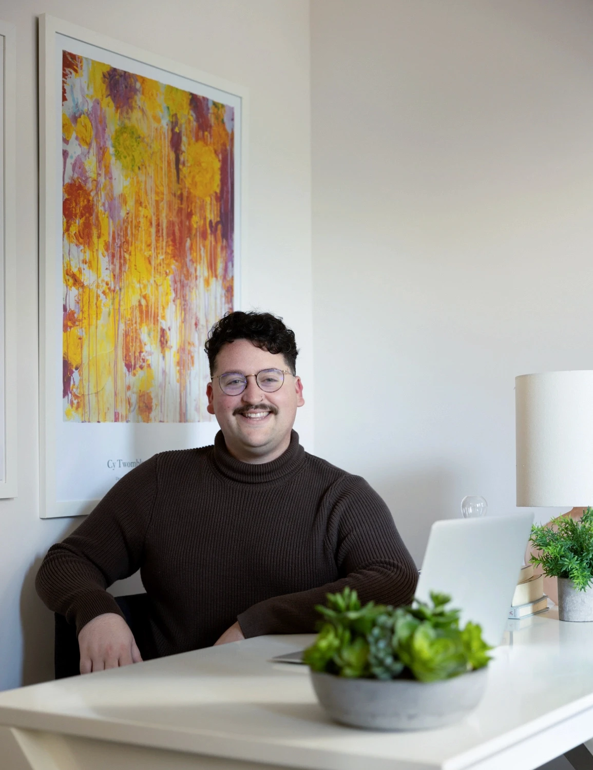 a queer expansive therapy therapist sitting on a chaiin new york city office in front of a vibrant orange and red abstract painting painting representing exclusivity and support around connecting to your identity in LGBTQ therapy.