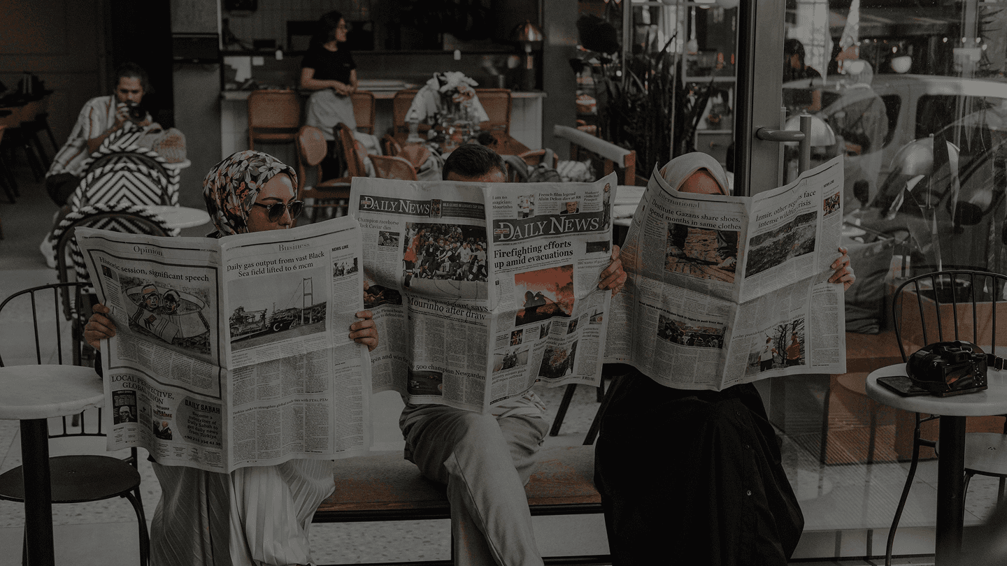 Several people sitting at a café table reading newspapers, with the papers covering most of their faces.