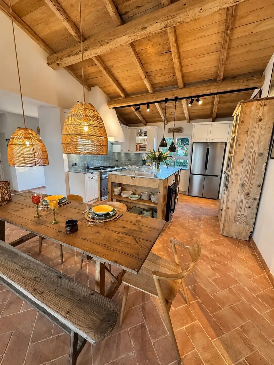 Vue d’ensemble de la cuisine de San Damiano avec îlot en bois, table à manger, sol en terre cuite et charpente apparente