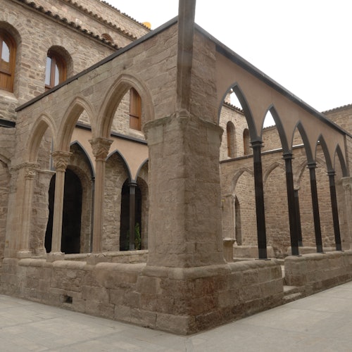 Stone courtyard with archways, pillars, and a surrounding building. Brown arched windows are visible above the courtyard.