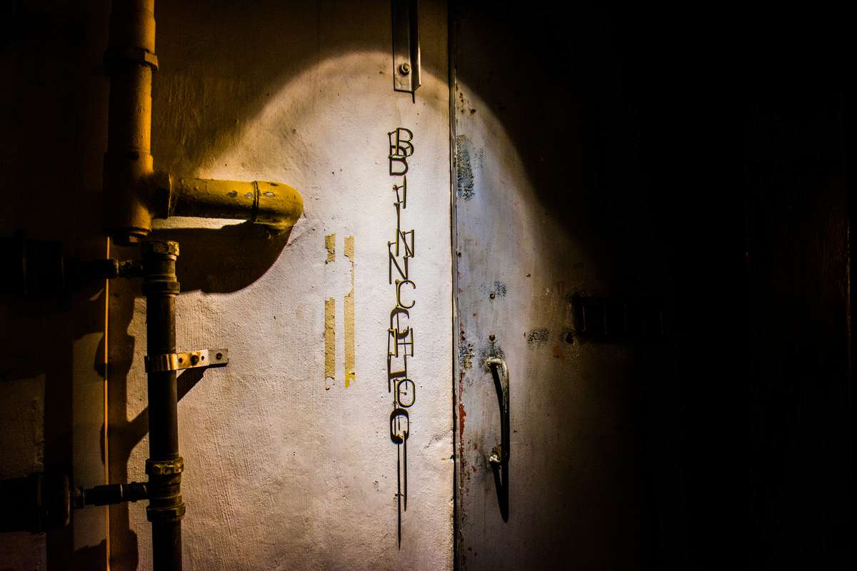 A spotlight illuminates graffiti on a textured wall next to industrial pipes and a metal door in a dark corridor.