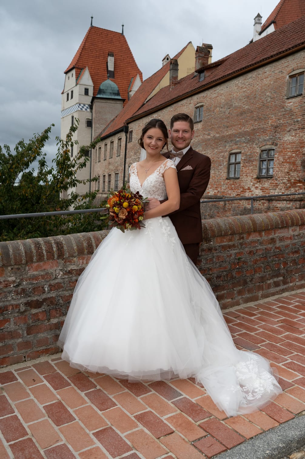 Kundenfeedback Hochzeitspaar für Maria Wiethaler freie Rednerin und Sängerin. Glückliches Brautpaar vor historischer Kulisse: Die Braut trägt ein elegantes Spitzen-Hochzeitskleid mit weitem Tüllrock und hält einen herbstlichen Blumenstrauß in warmen Rot- und Orangetönen. Der Bräutigam steht hinter ihr in einem stilvollen braunen Anzug mit passender Fliege. Beide lächeln glücklich in die Kamera, während sie auf einer alten Burgmauer mit Backsteinboden posieren. Hochzeitsfotografie mit romantischer, traditioneller Atmosphäre.
