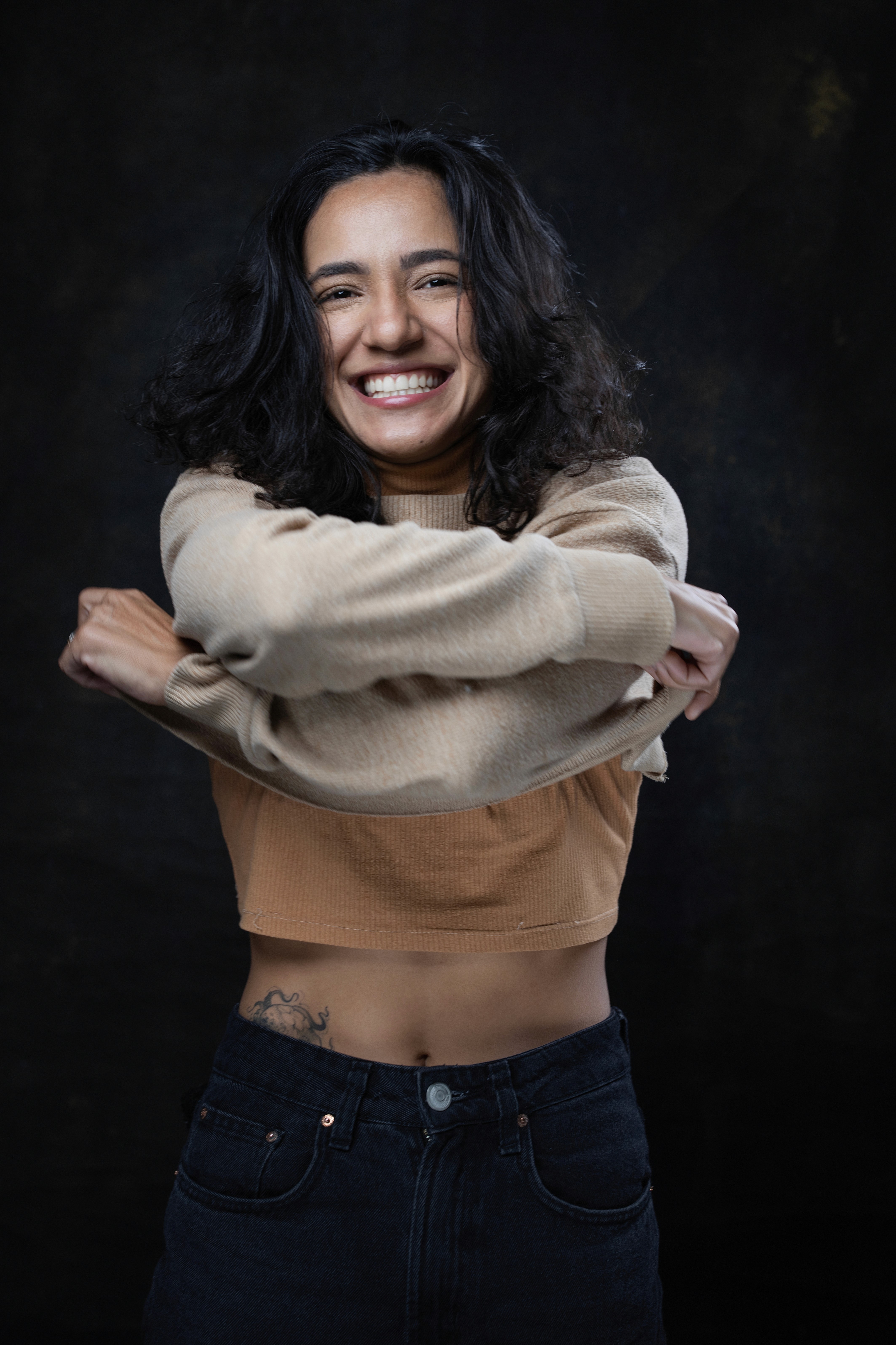 Una persona con una camiseta negra y pantalones de color claro está de pie en la cima rocosa de una montaña al atardecer, sosteniendo una cámara frente a su cara para tomar una foto. Un mar de nubes se extiende detrás de ellos, iluminado por la luz dorada del sol poniente.