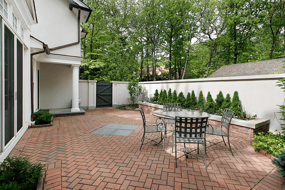 A formal brick patio with a small metal dining table and chairs, bordered by a low retaining wall with neatly trimmed greenery. The space is surrounded by a white wall and mature trees, providing a secluded, peaceful atmosphere.