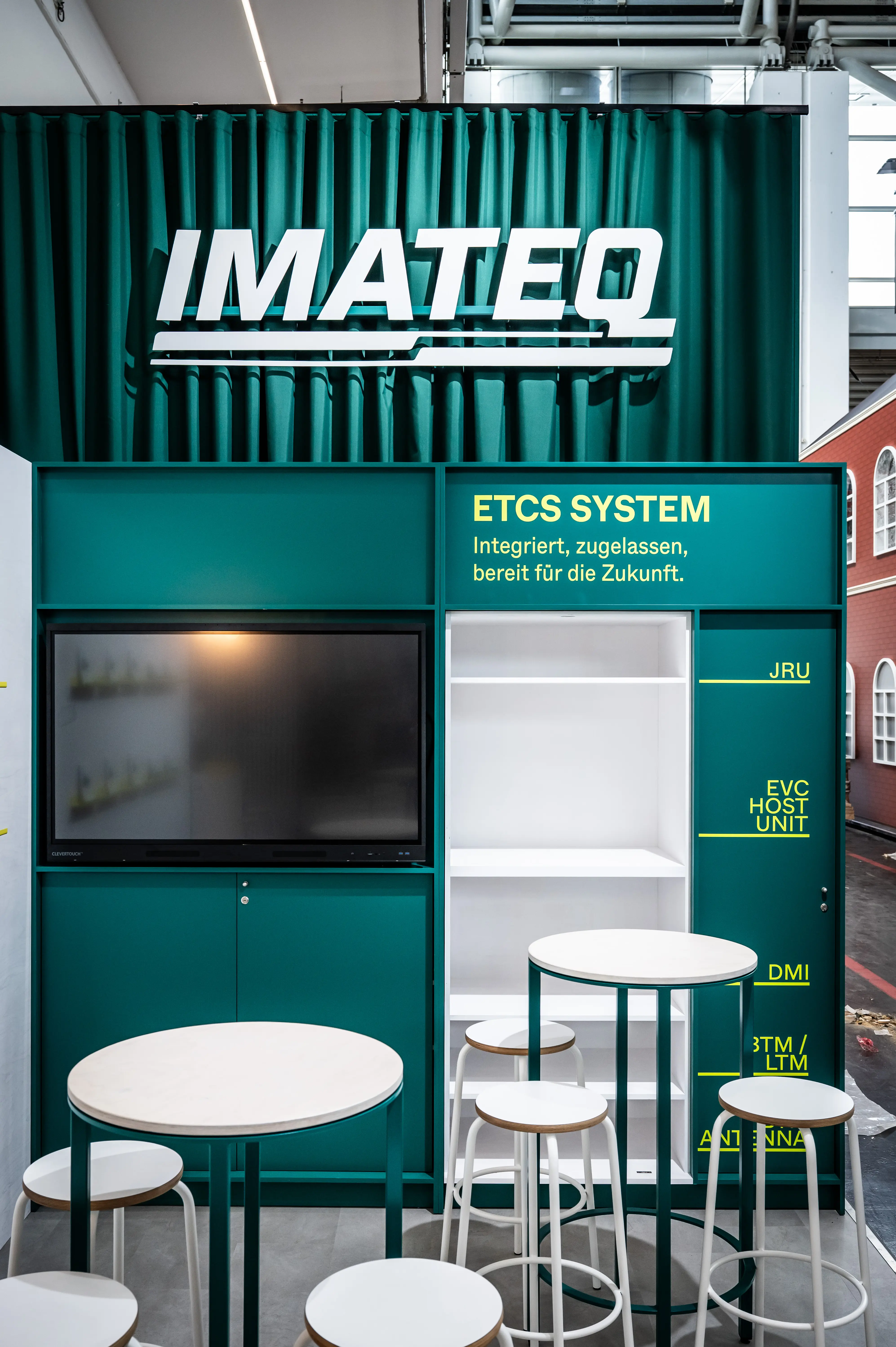 Green Imateq wall with ETCS system display, screen, white shelves and bar stools at the booth.