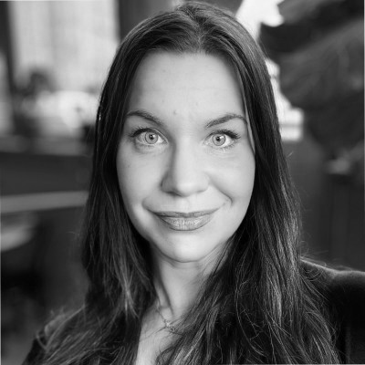 A smiling person with curly hair, wearing a leather jacket, stands in a cozy indoor setting.