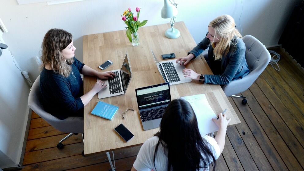 Drie vrouwen werken samen rond een tafel met laptops en schrijfwaren in een heldere, moderne werkruimte.
