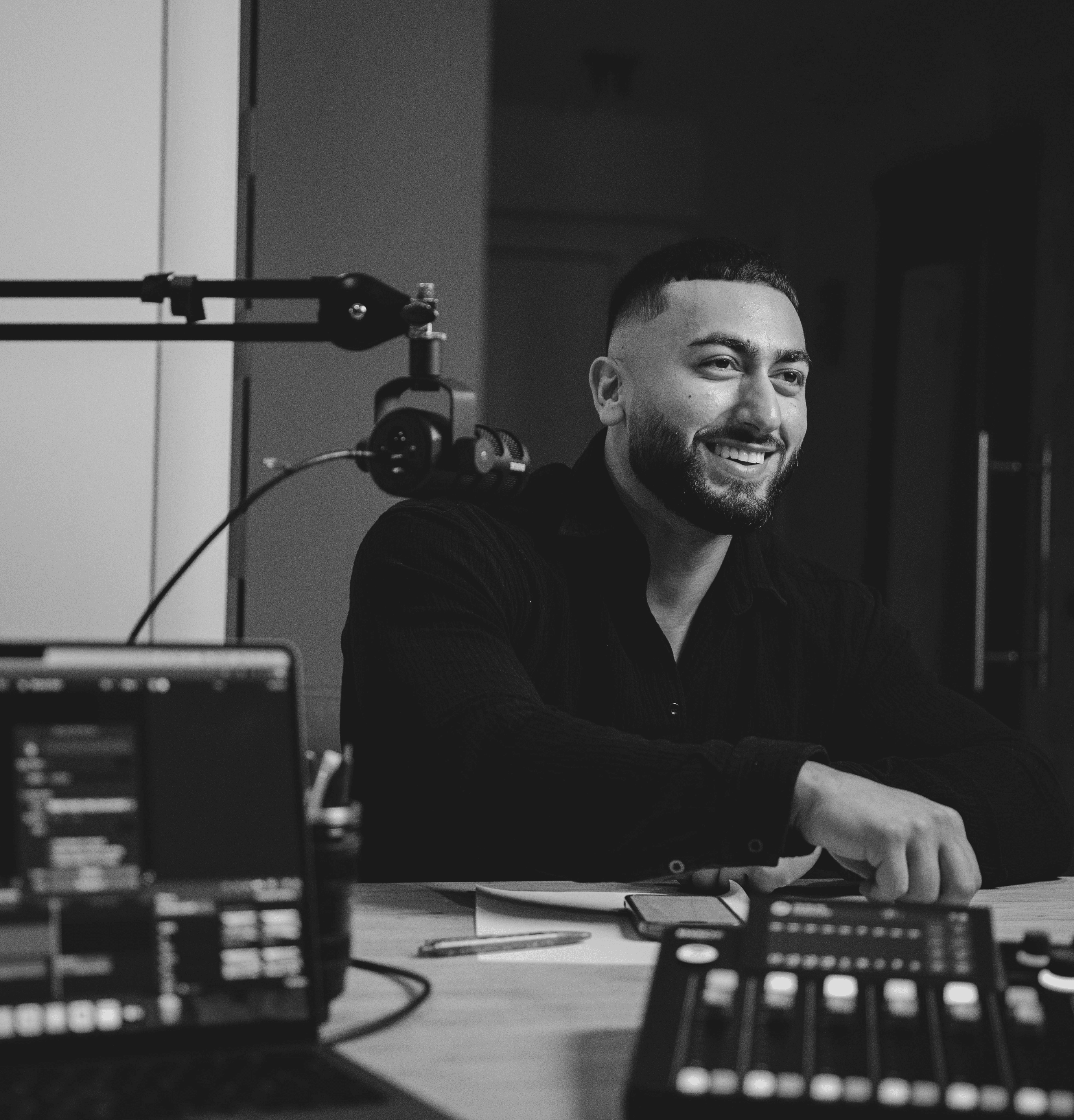 Black and white photo of FitbyElyas sitting at a desk with audio equipment and smiling on a podcast.