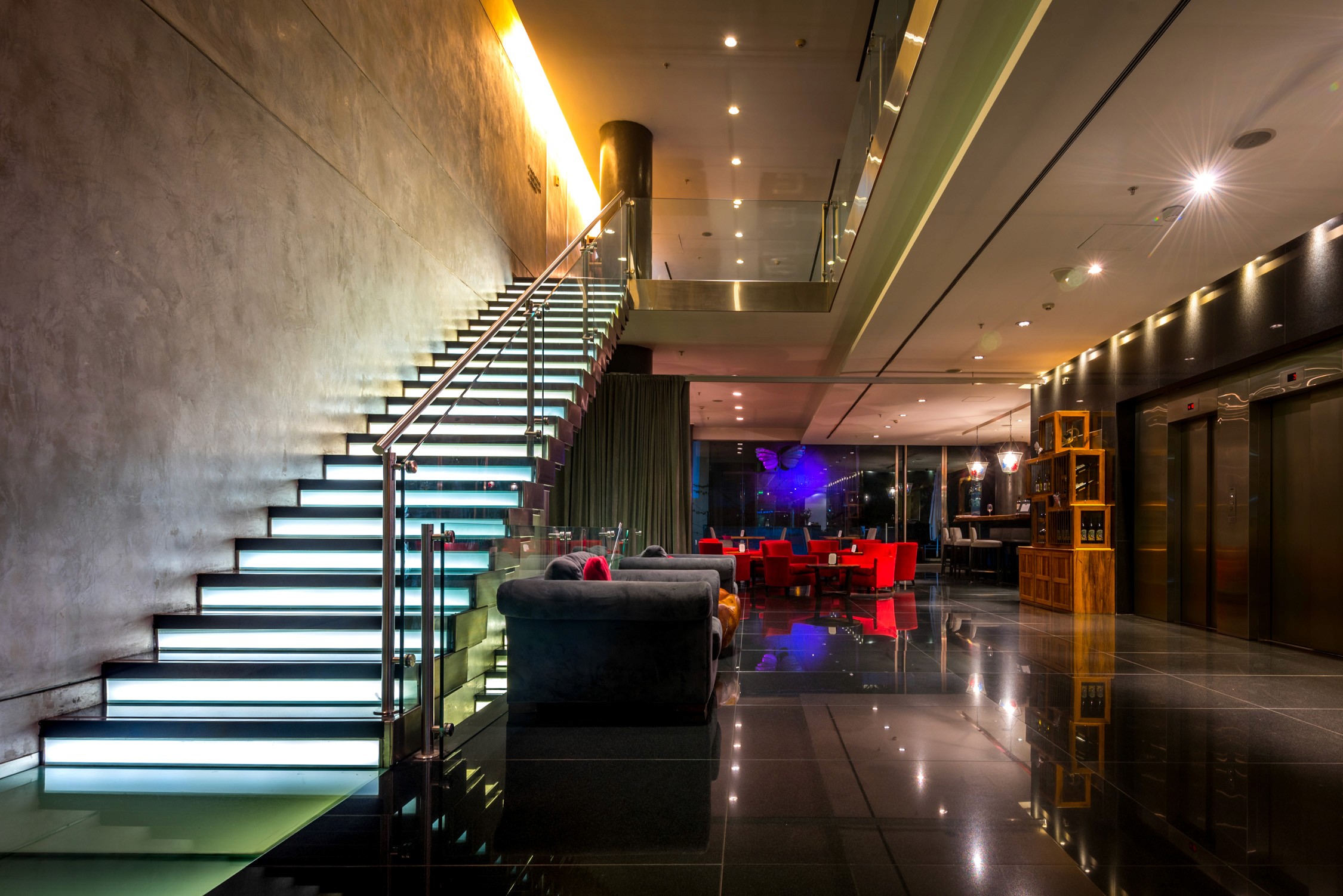 A modern lounge with glowing white stairs, a sleek reception desk, and a dimly lit bar area with neon accents.