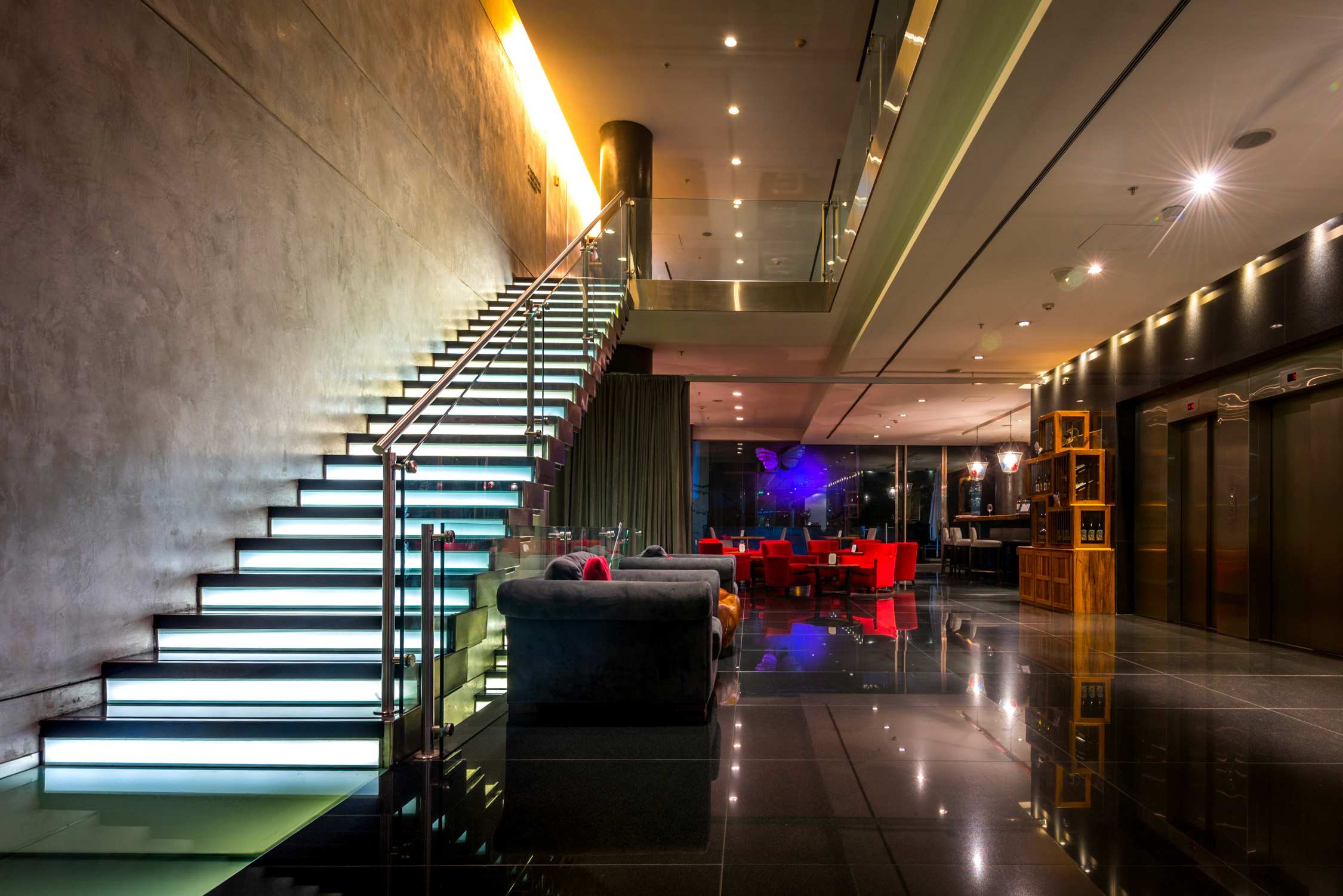 A modern lounge with glowing white stairs, a sleek reception desk, and a dimly lit bar area with neon accents.