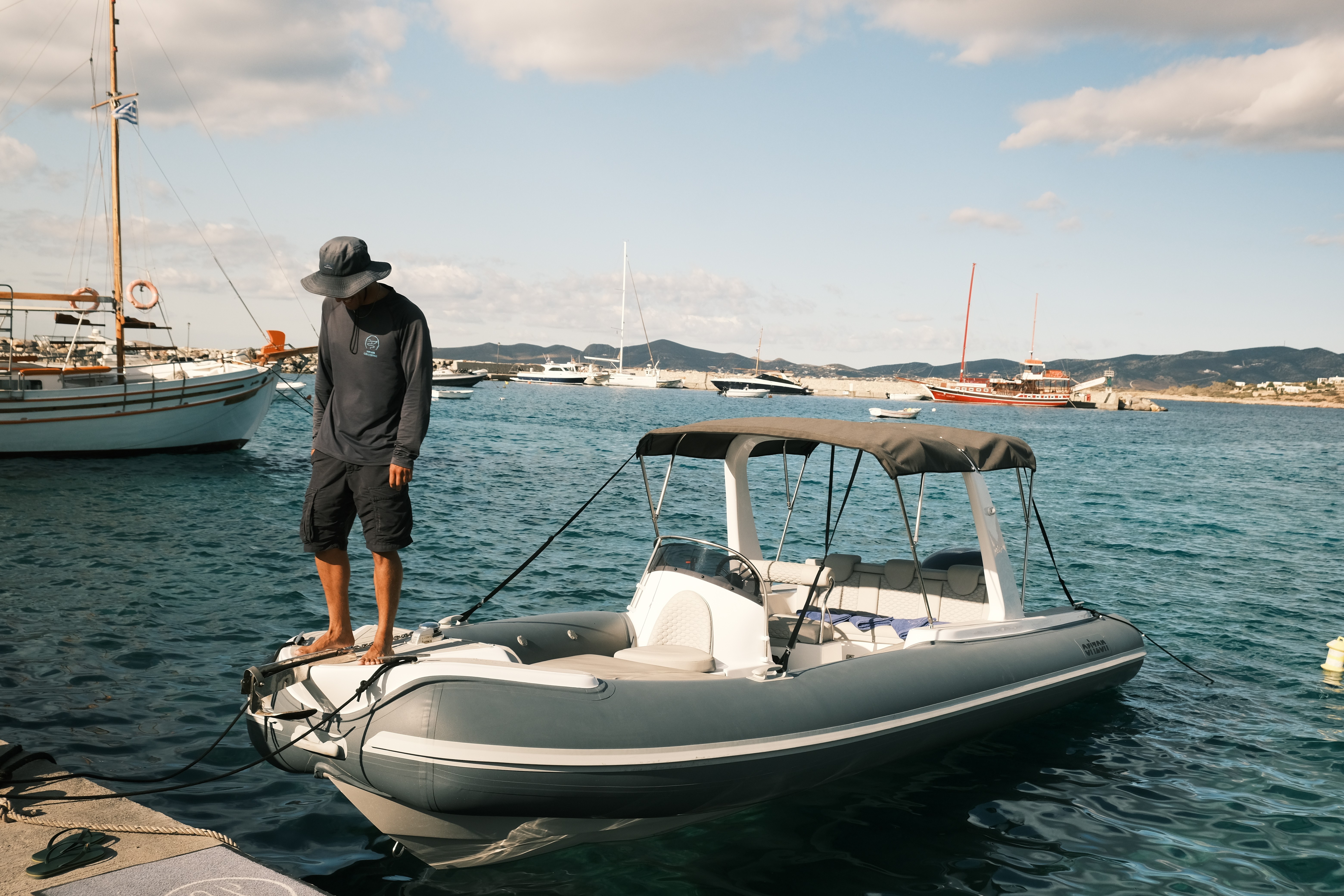 Crew member standing on the bow of a gray and white rigid inflatable boat in calm turquoise waters with traditional Greek sailboats and hills in background.