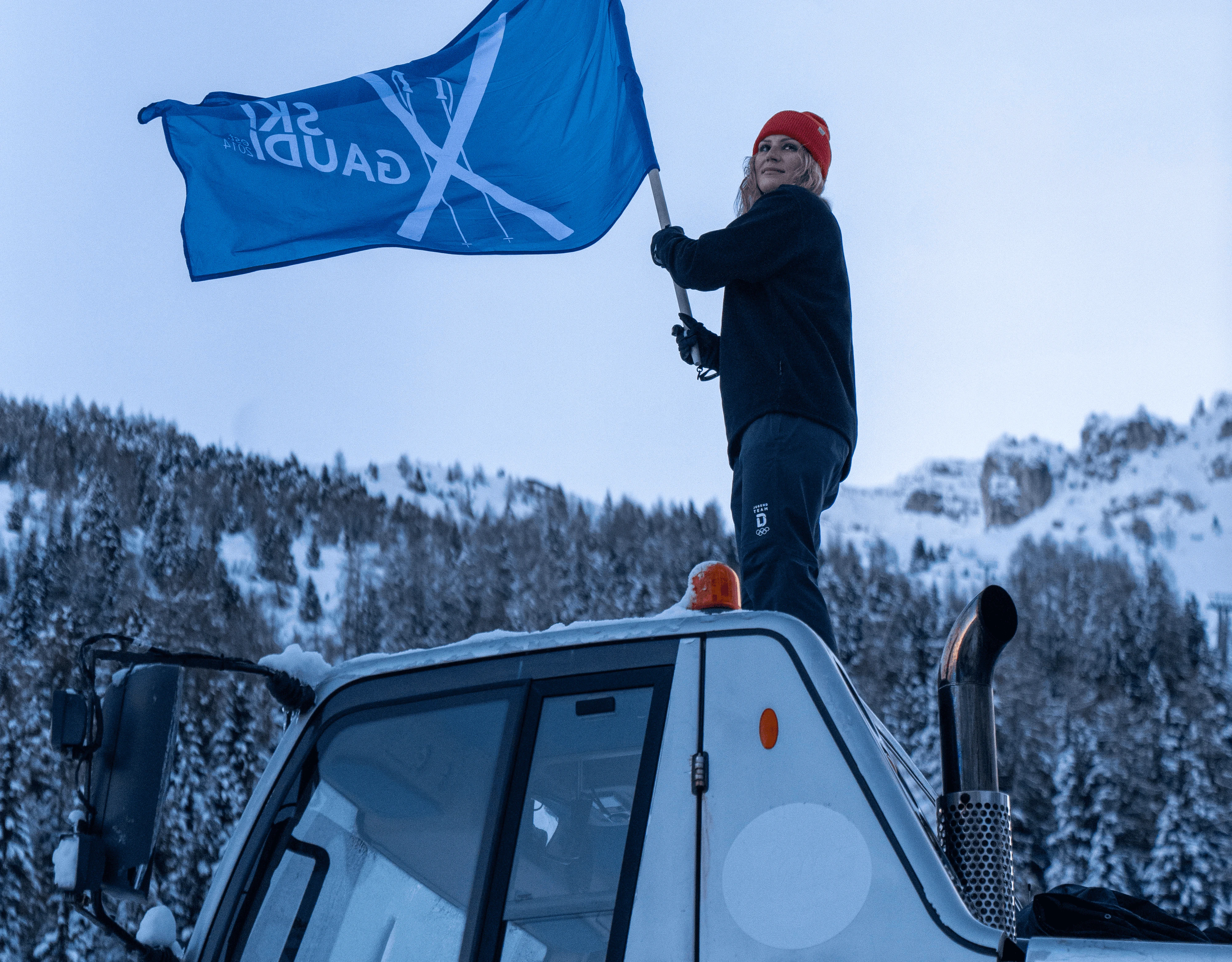 Person waving blue flag on snowcat in snowy mountain landscape.
