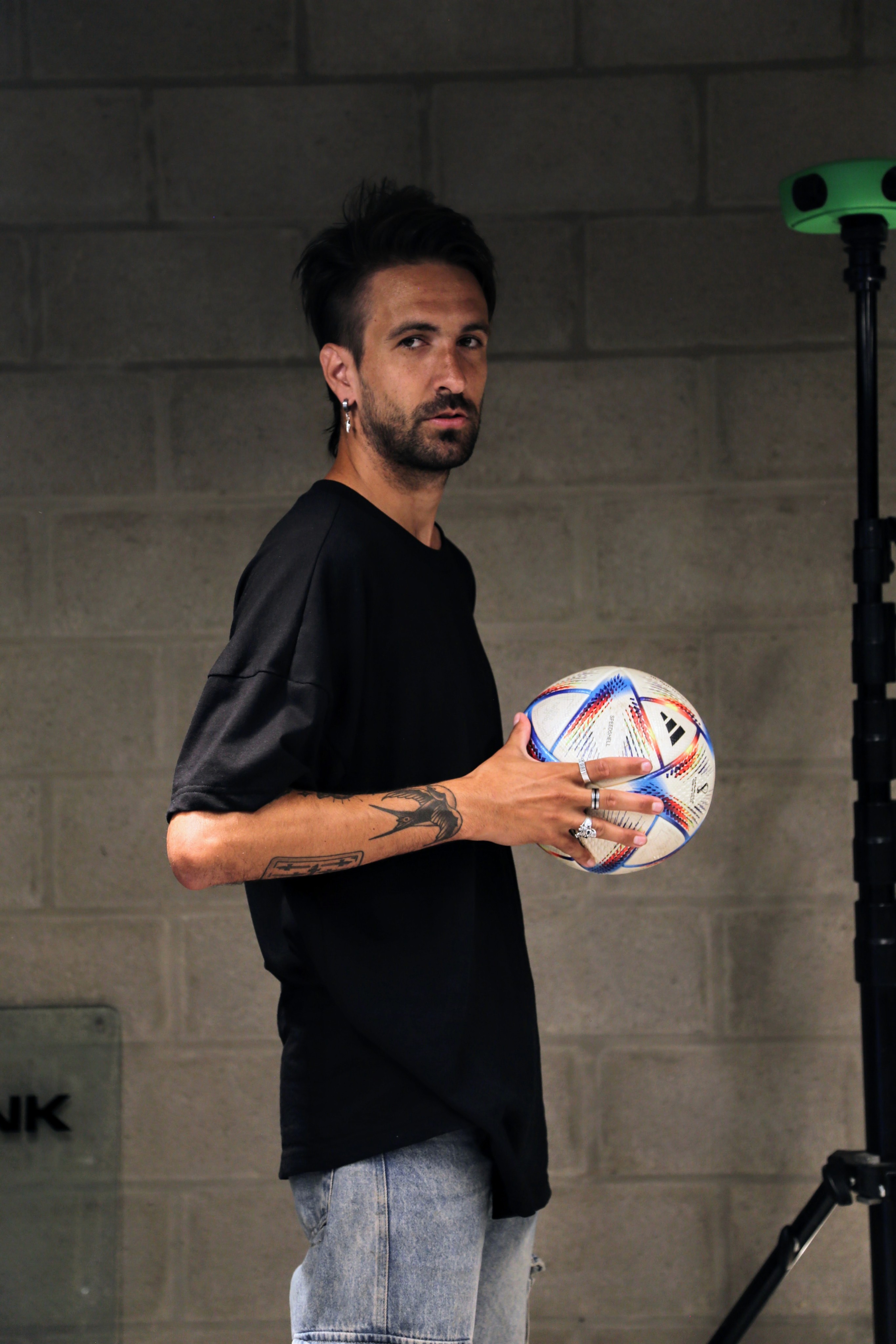A man in a blue bomber jacket stands against a dark background. The lighting is dramatic, highlighting his focused expression. His pose is confident.