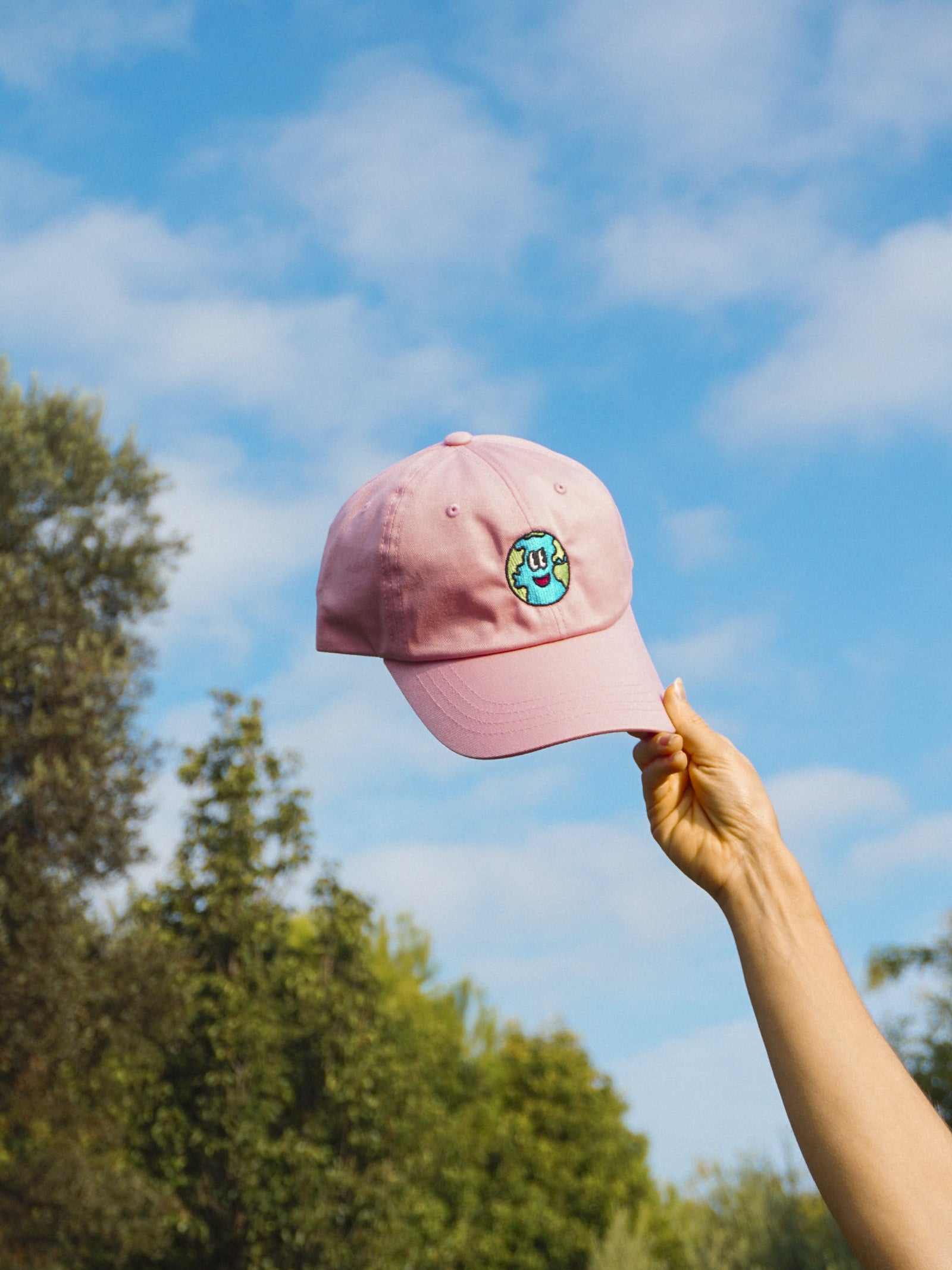 Pink cap with a logo held up against a blue sky with trees in the background