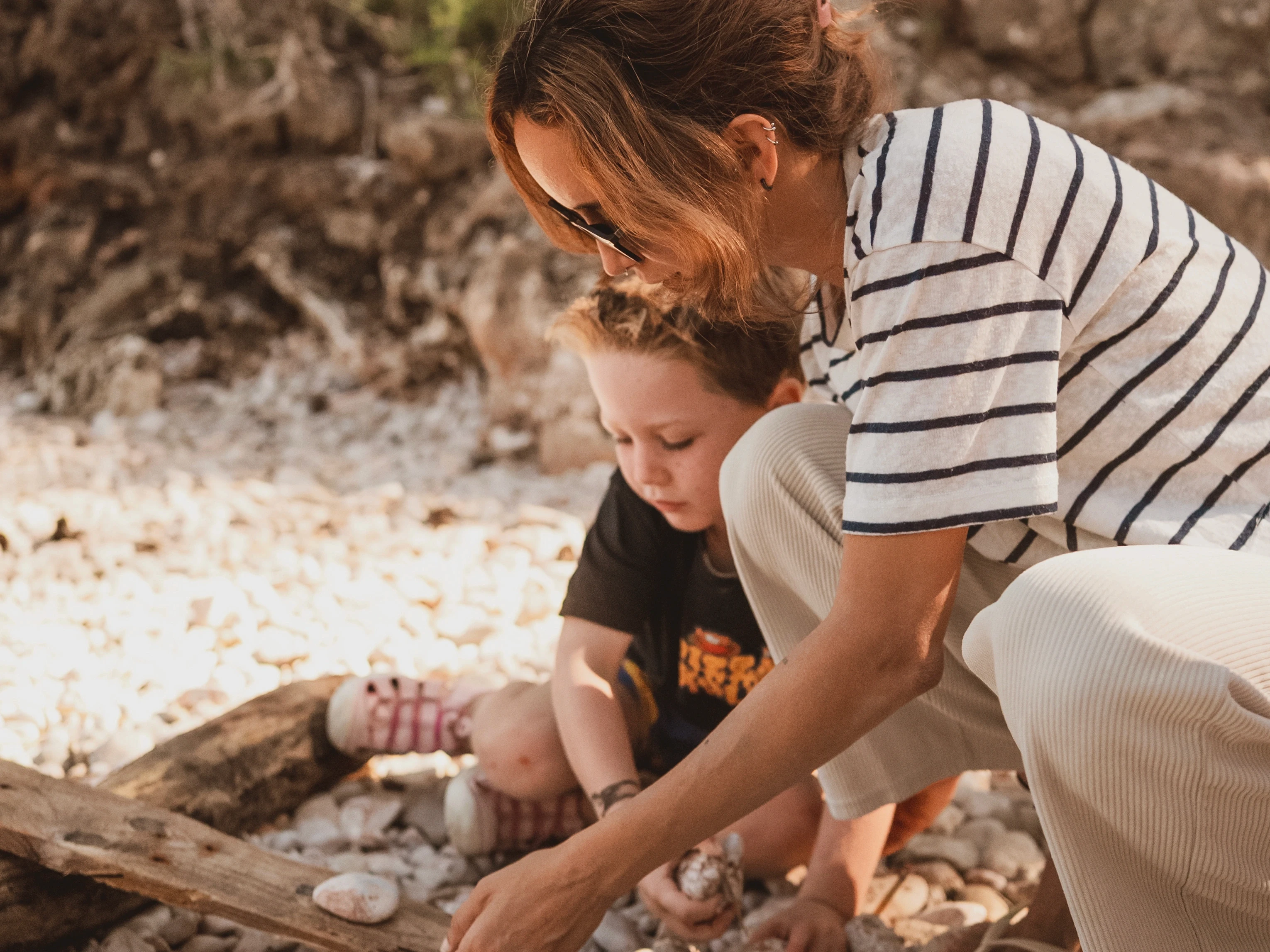 Close-up of an mother and young child crouching on a rocky beach, arranging driftwood and picking up stones together.