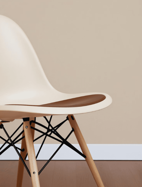 A minimalist chair with a cream shell, brown seat cushion, and wooden legs connected by black metal rods against a beige wall and wood floor background.