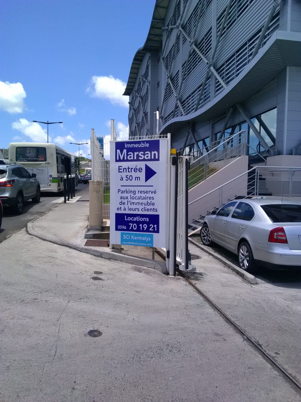 Entrée d'un parking à côté d'un bâtiment moderne avec une façade incurvée et des poutres métalliques. Un panneau indique « Immeuble Marsan » en français. Des voitures et un bus se trouvent à proximité sous un ciel bleu.