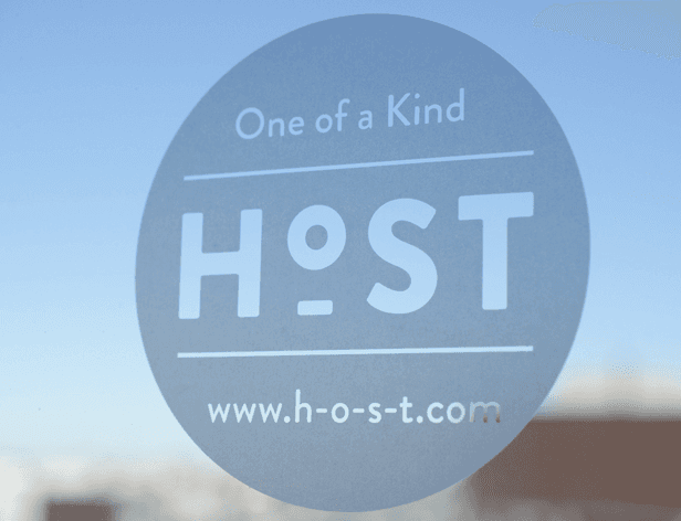 The image shows a circular sticker with a muted gray background and white text. The text reads "One of a Kind" above the word "H9ST", and "www.h-o-s-t.com" is below it. The sticker is positioned slightly off-center on a reflective surface, with a blurred blue sky and a hint of a brown/beige object visible in the out-of-focus background.