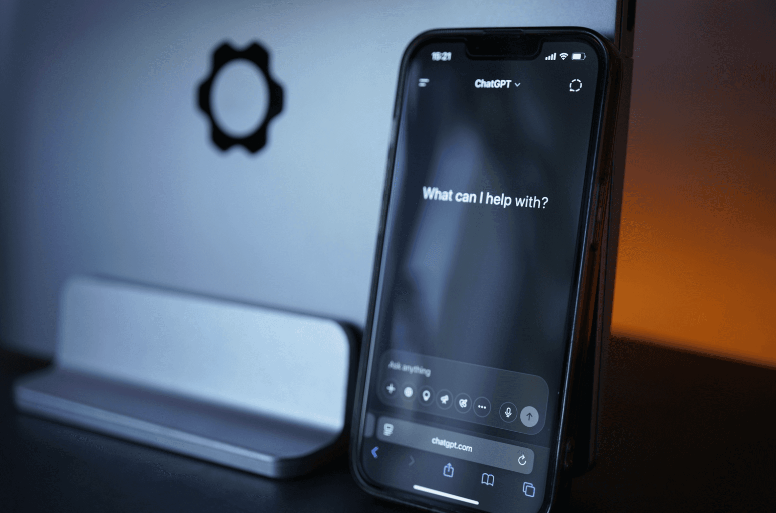 A smartphone displaying the ChatGPT app in dark mode with the prompt “What can I help with?” shown on screen, leaning against a laptop on a dimly lit desk with cool blue and warm orange lighting in the background.