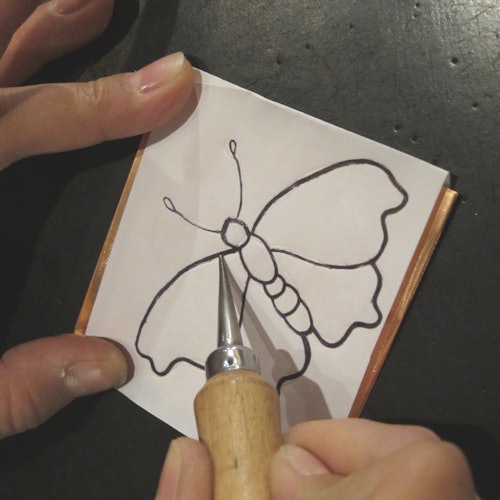 Hai bàn tay giữ tờ giấy có hình con bướm, đang được chạm khắc bằng một công cụ.