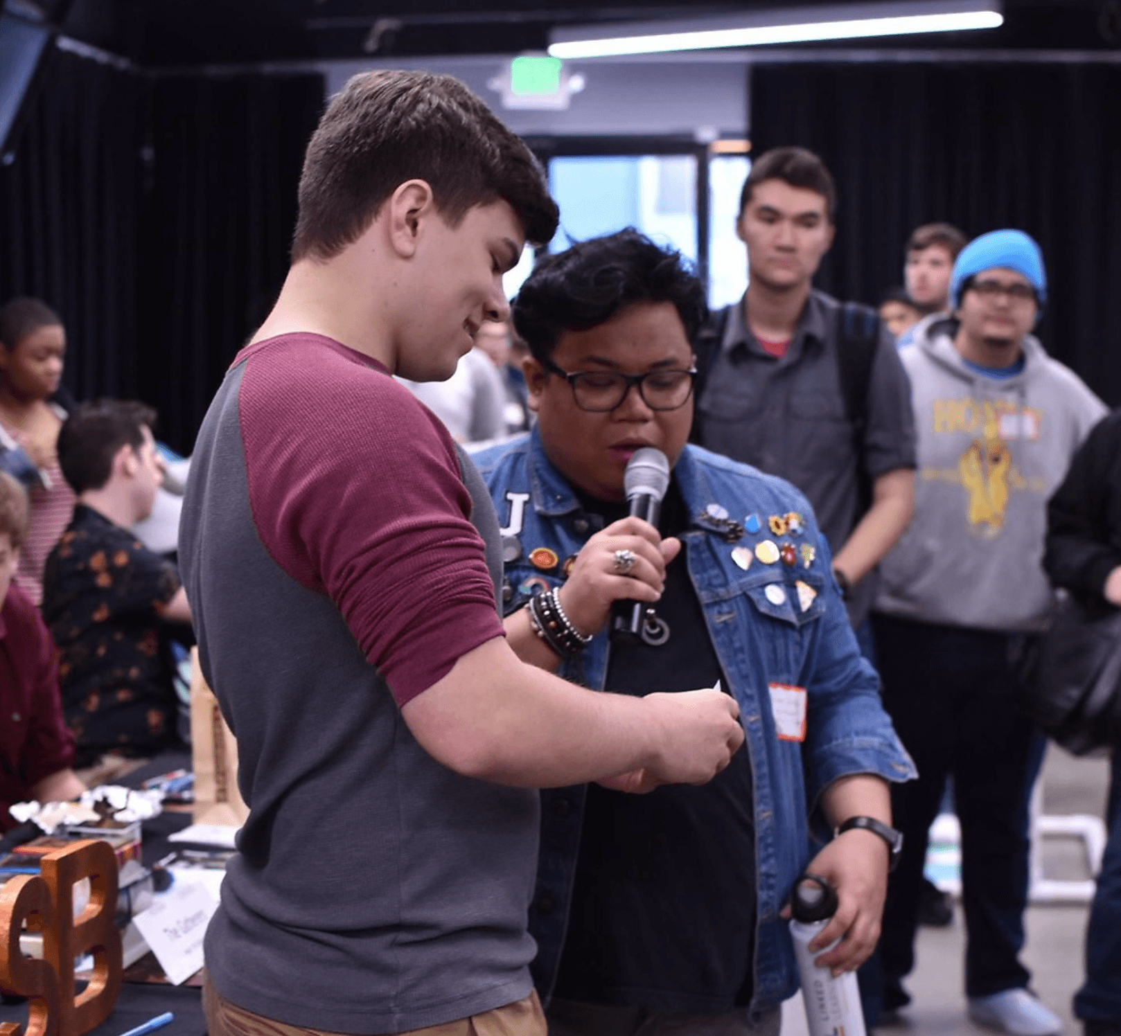 A diverse group of young adults, some wearing casual clothing and others with jackets adorned with colorful pins, engage in a lively exchange at an indoor event, highlighted by a speaker using a microphone.
