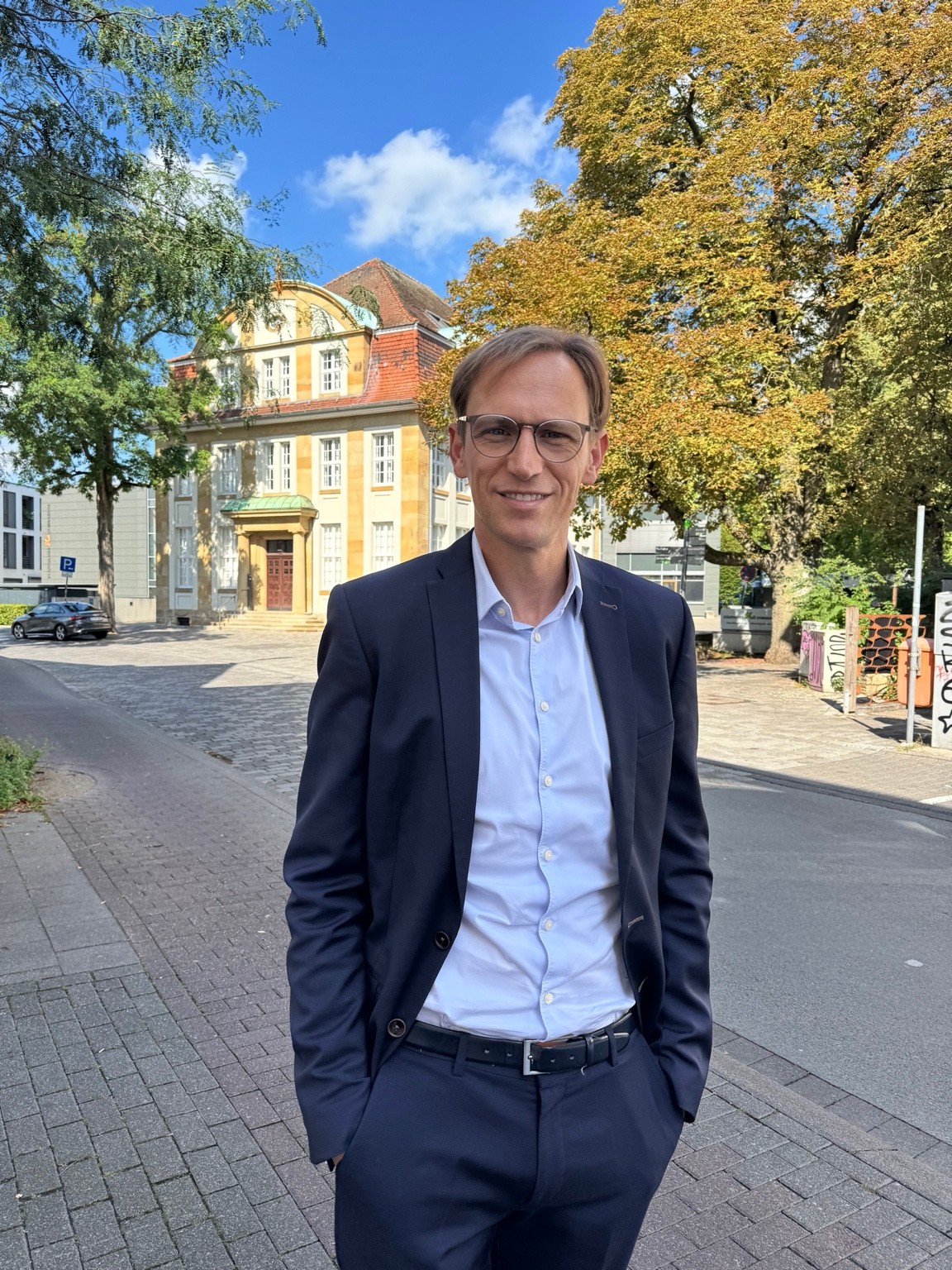 Dominik Minkus im Freien, im dunkelblauen Anzug und mit offenem Hemdkragen.